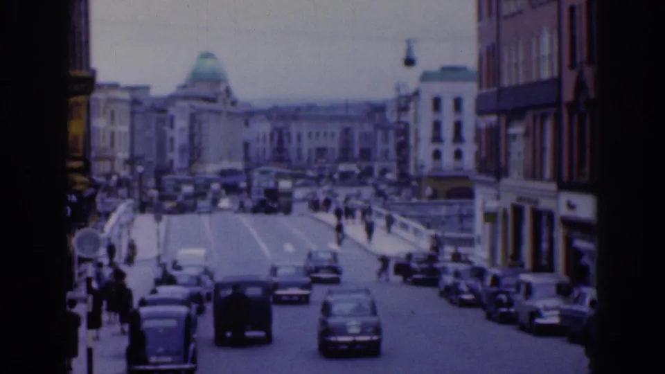 1961 Dublin city street with moving cars, parked vehicles along the curb, pedestrians near the roadway, and a domed building visible beyond the traffic.