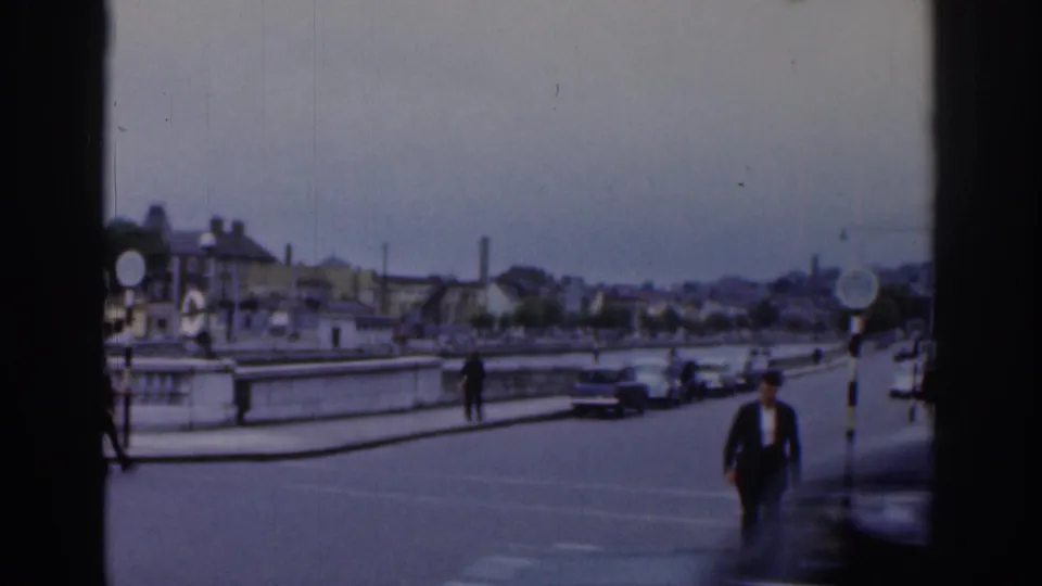 1961 Dublin riverside street view with bridge or quay railings, cars passing through the frame, pedestrians crossing or walking nearby, and low buildings beyond the roadway.
