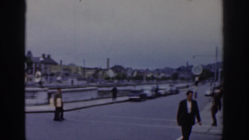 1961 Dublin riverside roadway with pedestrians in mid-century clothing, cars moving across the street, bridge or quay railings, and distant city buildings under a cloudy sky.