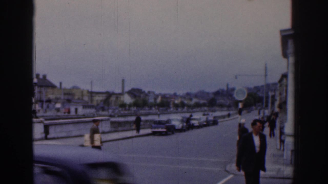 1961 Dublin street scene with pedestrians near a riverside crossing, parked cars, quay walls, and buildings under hazy light.