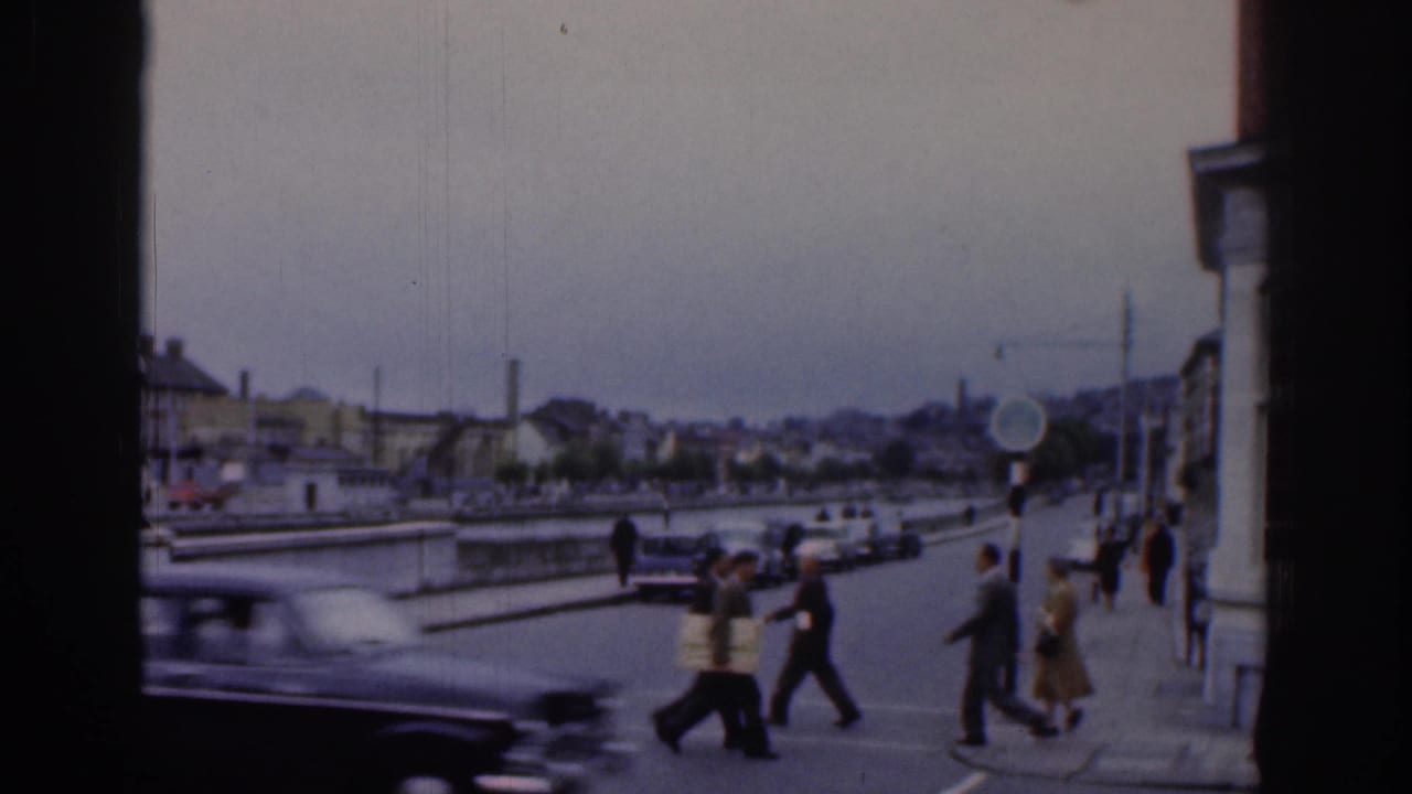 People cross a street as a dark car passes near the river.