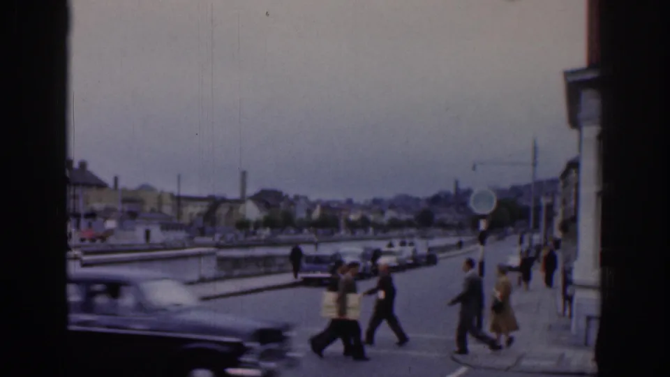 1961 Dublin pedestrians cross near a quay while a dark car blurs past and more people walk along the sidewalk.