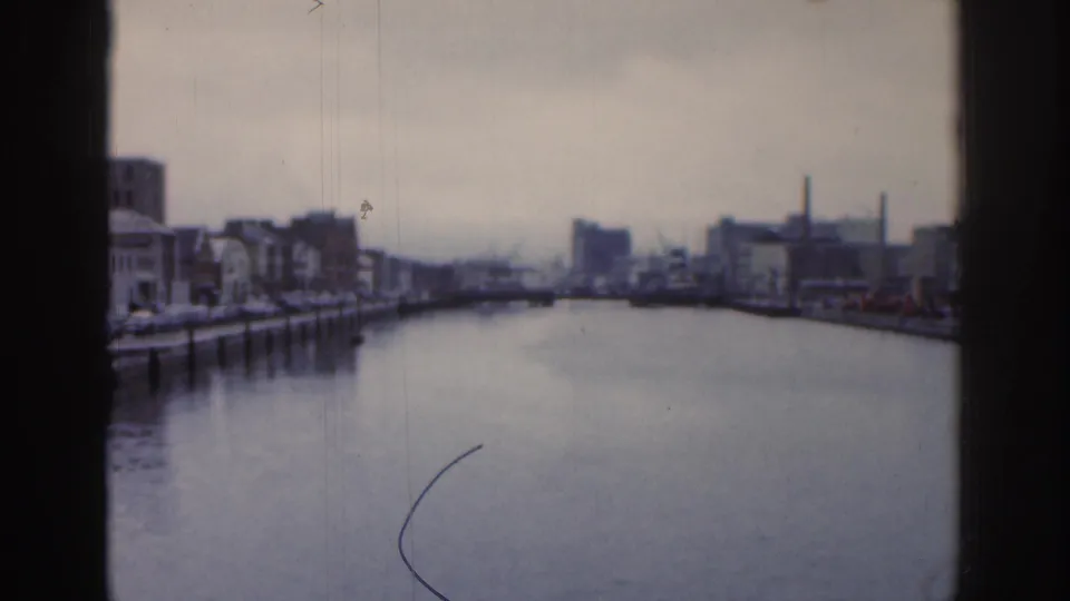 Wide 1961 Dublin river or canal view with quay walls, industrial buildings, smokestacks, and a bridge in the distance.