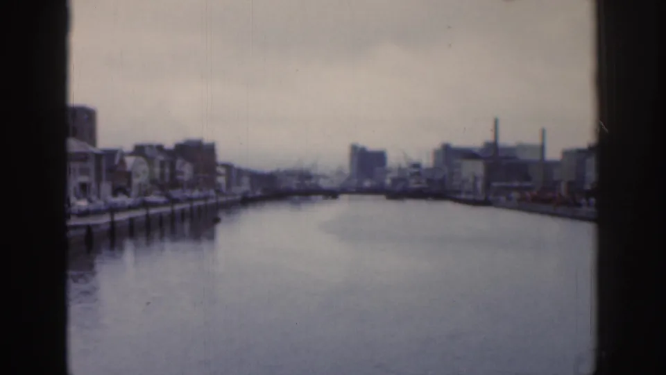 1961 Dublin waterfront panorama showing still water, quay-side buildings, smokestacks, and a low bridge under an overcast sky.