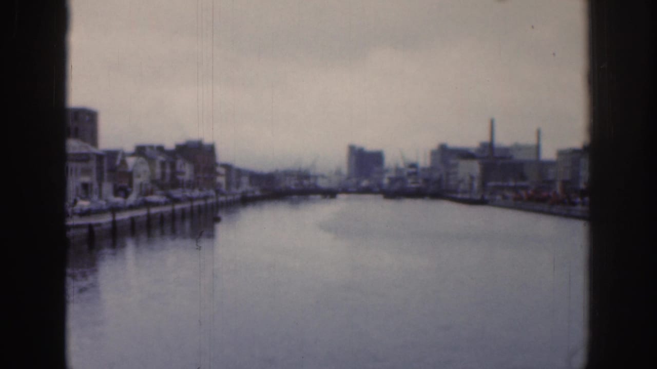 1961 Dublin waterway view through a window, with low buildings, quay edges, chimneys, and an overcast sky in soft 8mm focus.