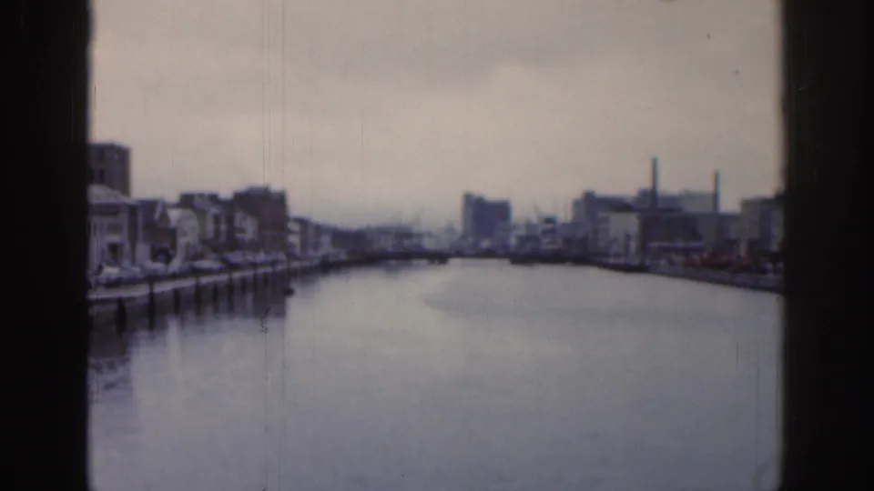 1961 Dublin waterway view through a window, with low buildings, quay edges, chimneys, and an overcast sky in soft 8mm focus.
