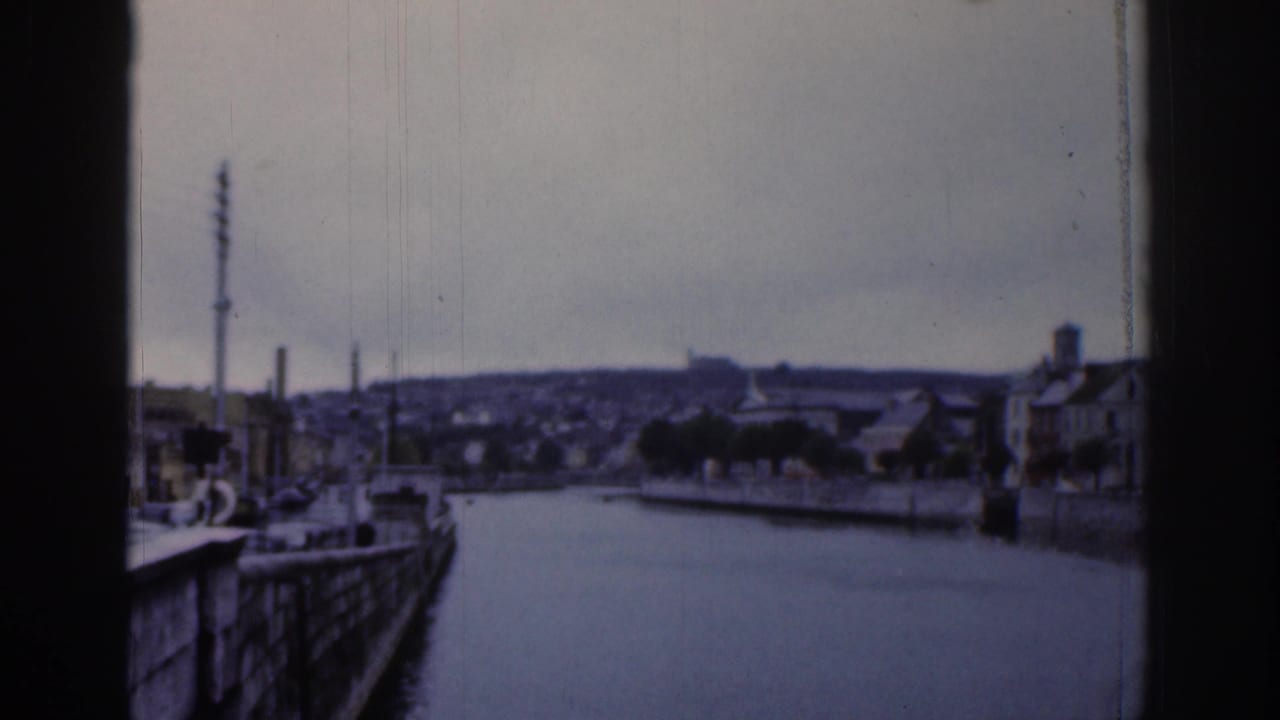 Waterway bend with quayside structures, rooftops, and a tall mast in the distance.
