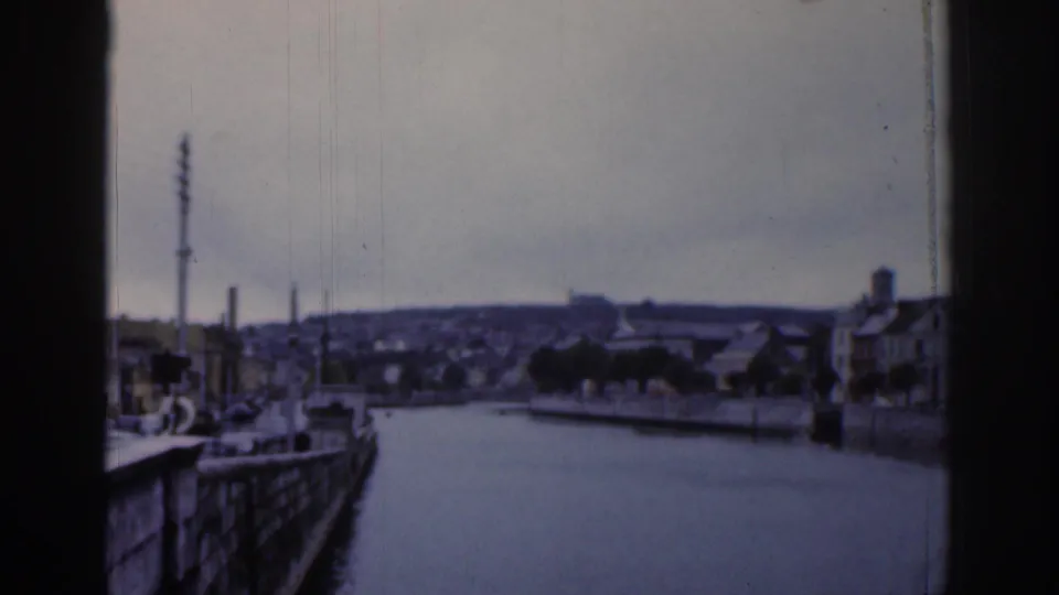 Dublin waterfront scene showing a calm channel, quayside edge, low rooftops, trees, and a tall mast-like structure under cloud cover.