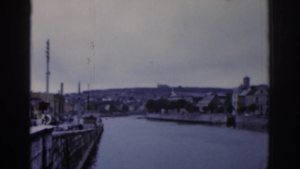 1961 Dublin home-movie frame of a calm waterfront, with quay walls, trees, rooftops, chimneys, and distant industrial silhouettes.