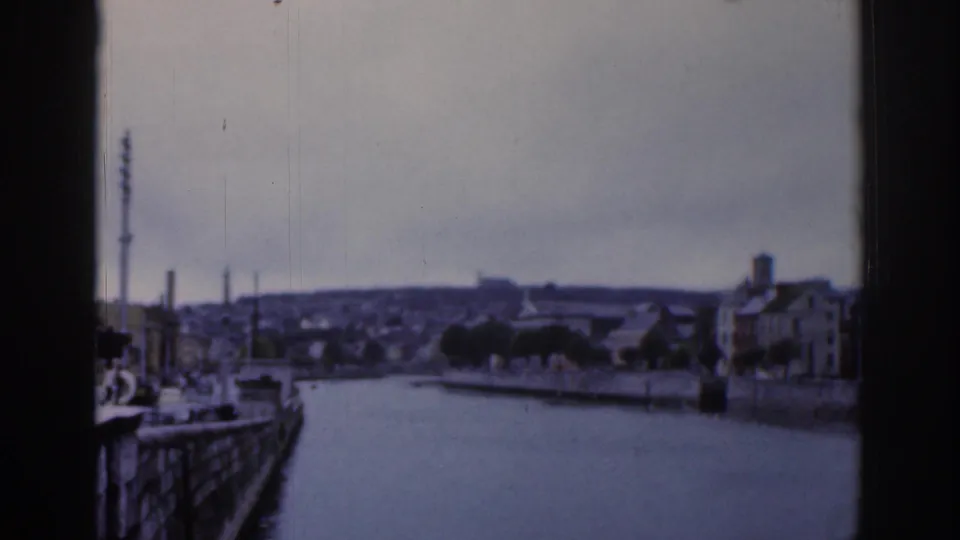 Dublin waterway seen through a dark window mullion, with quay walls, trees, low buildings, a tall mast, and muted 8mm color.