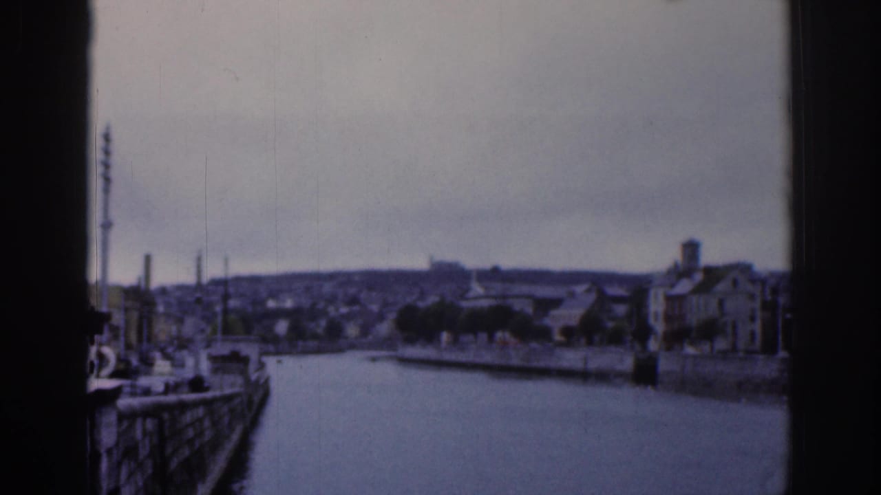 1961 Dublin 8mm view of a wide urban waterway, stone quay walls, moored boats, and distant hillside buildings seen through dark frame edges.