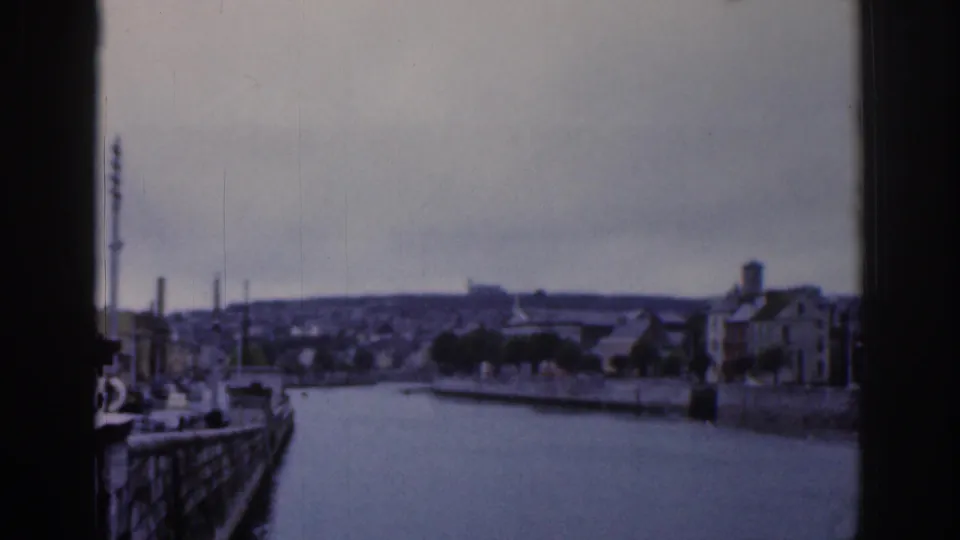 1961 Dublin 8mm view of a wide urban waterway, stone quay walls, moored boats, and distant hillside buildings seen through dark frame edges.