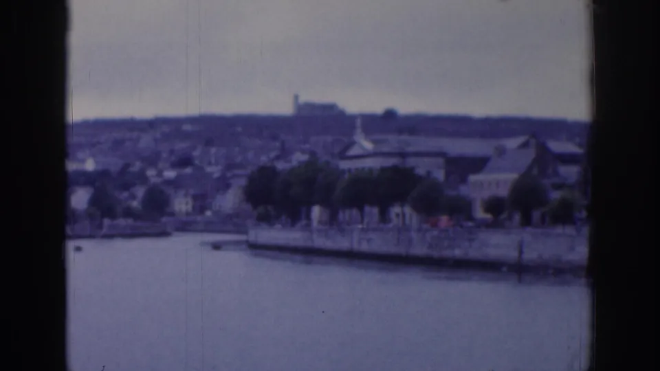 1961 Dublin waterway scene with a tree-lined stone bank, low buildings, hillside housing, and a distant spire-like structure under gray sky.