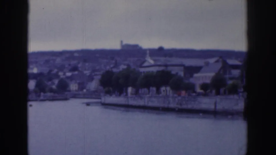 Dublin 1961 home-movie frame showing the waterway, quay wall, riverside trees, low buildings, and a hazy hillside skyline in faded color.