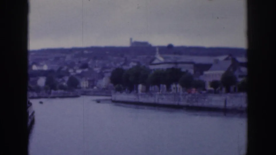 1961 Dublin 8mm frame of an urban waterside embankment, trees, low structures, and a distant hillside, with dark film-edge framing.
