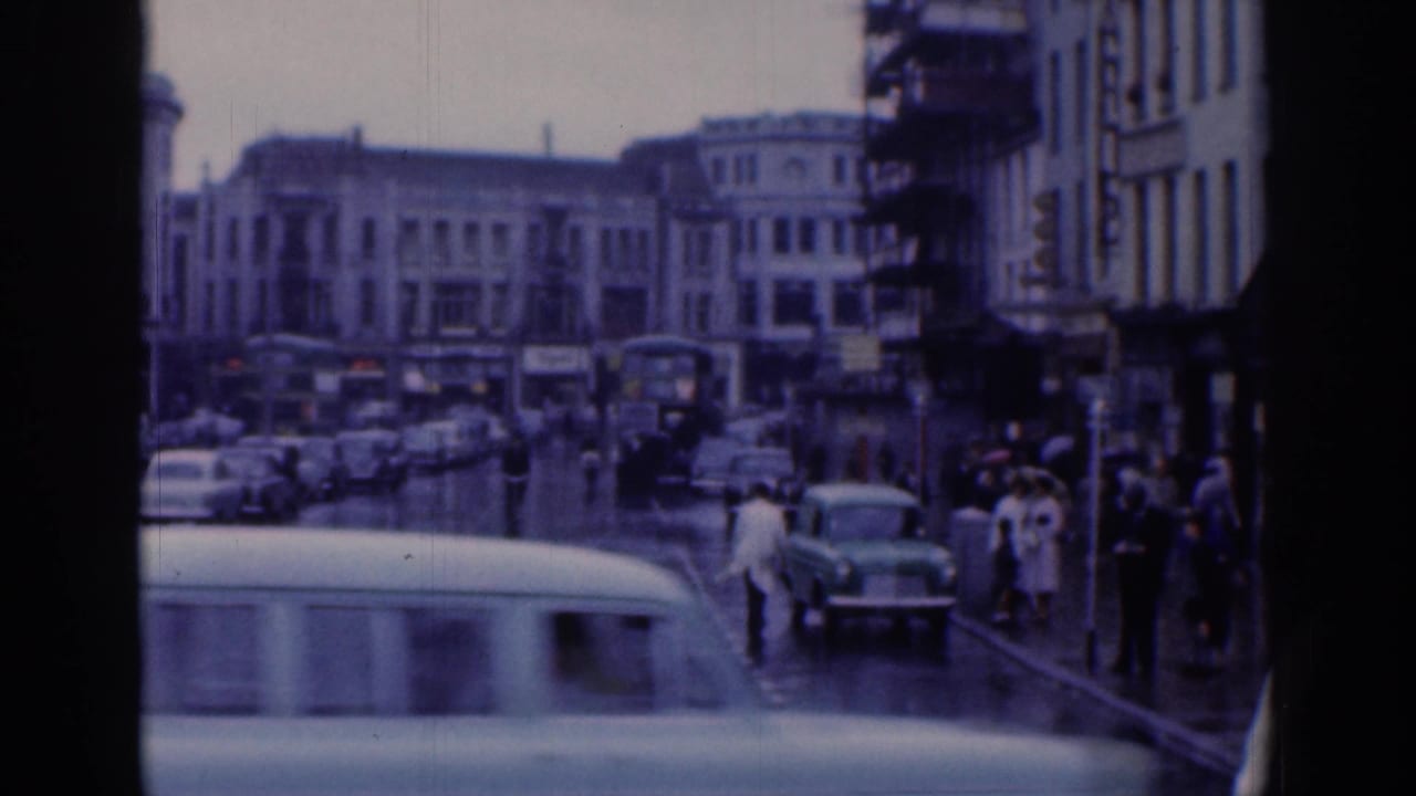 Rainy Dublin street scene with mid-century cars, pedestrians on sidewalks, umbrellas, and urban building fronts.