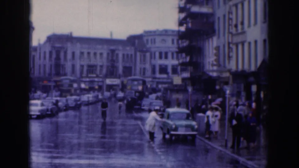 1961 Dublin street view showing rain-dark pavement, parked and moving cars, pedestrians, and period commercial buildings.