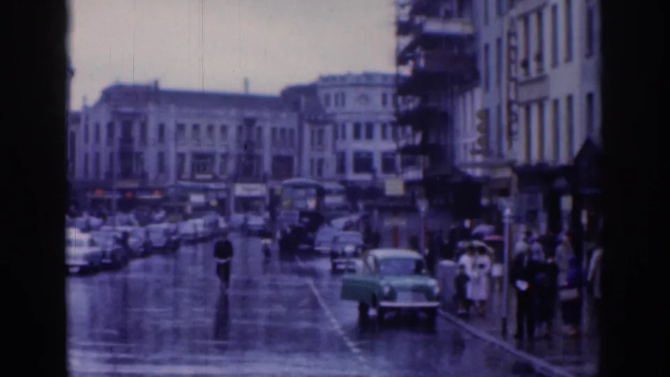 Soft 8mm view of a rainy Dublin street with a green car, pedestrians, umbrellas, traffic, and period architecture.