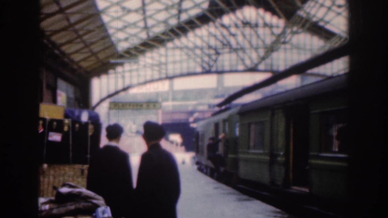 Two passengers in dark coats walk away from the camera on a covered Dublin railway platform in 1961, with a green passenger train stopped beside them.