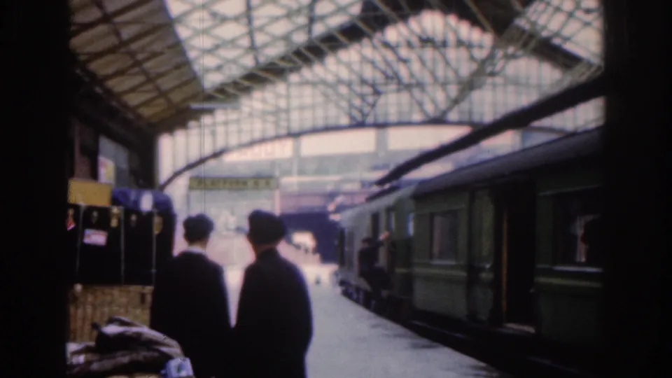 Two passengers in dark coats walk away from the camera on a covered Dublin railway platform in 1961, with a green passenger train stopped beside them.