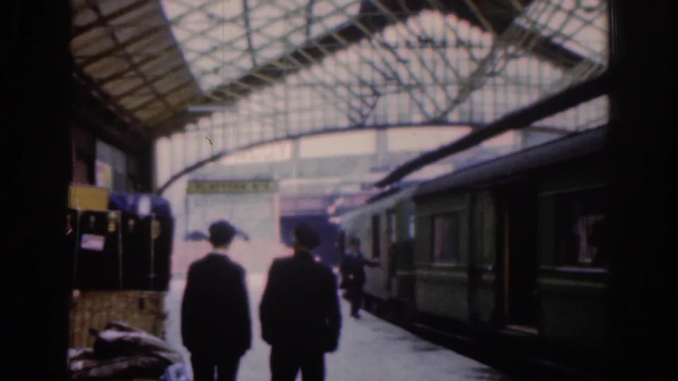 Two dark-clad passengers move along the Dublin station platform in 1961 as a green passenger train stands alongside the arched glass-and-steel roof.