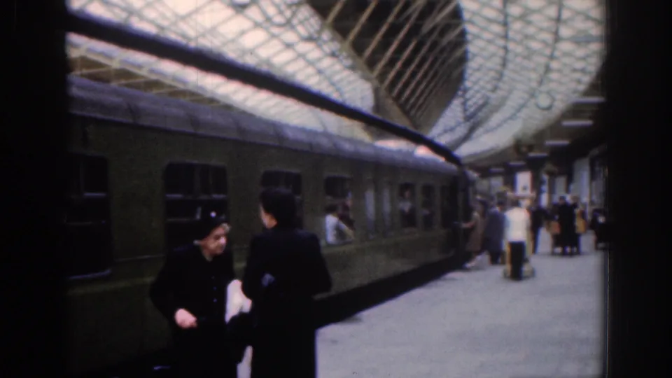 Several travelers stand or walk along a covered Dublin railway platform in 1961, with a green train carriage, visible windows, and luggage in the background.