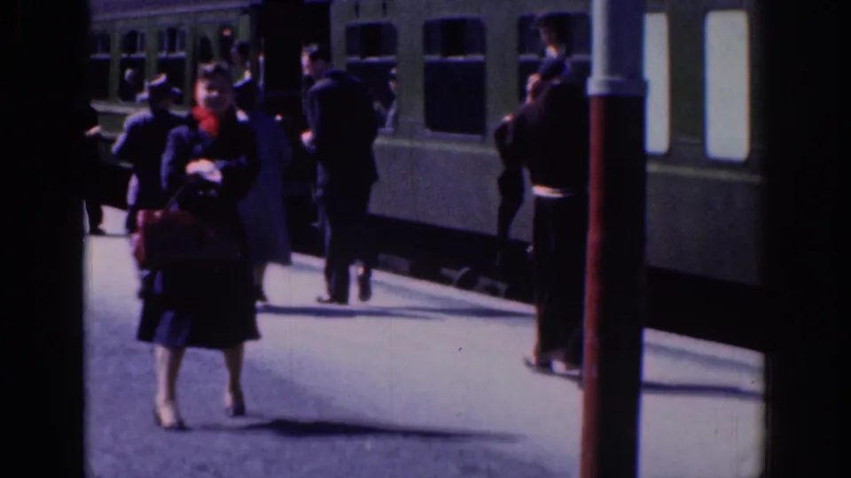 Passengers in period coats walk and stand beside a green train on a Dublin station platform in 1961 home-movie footage.