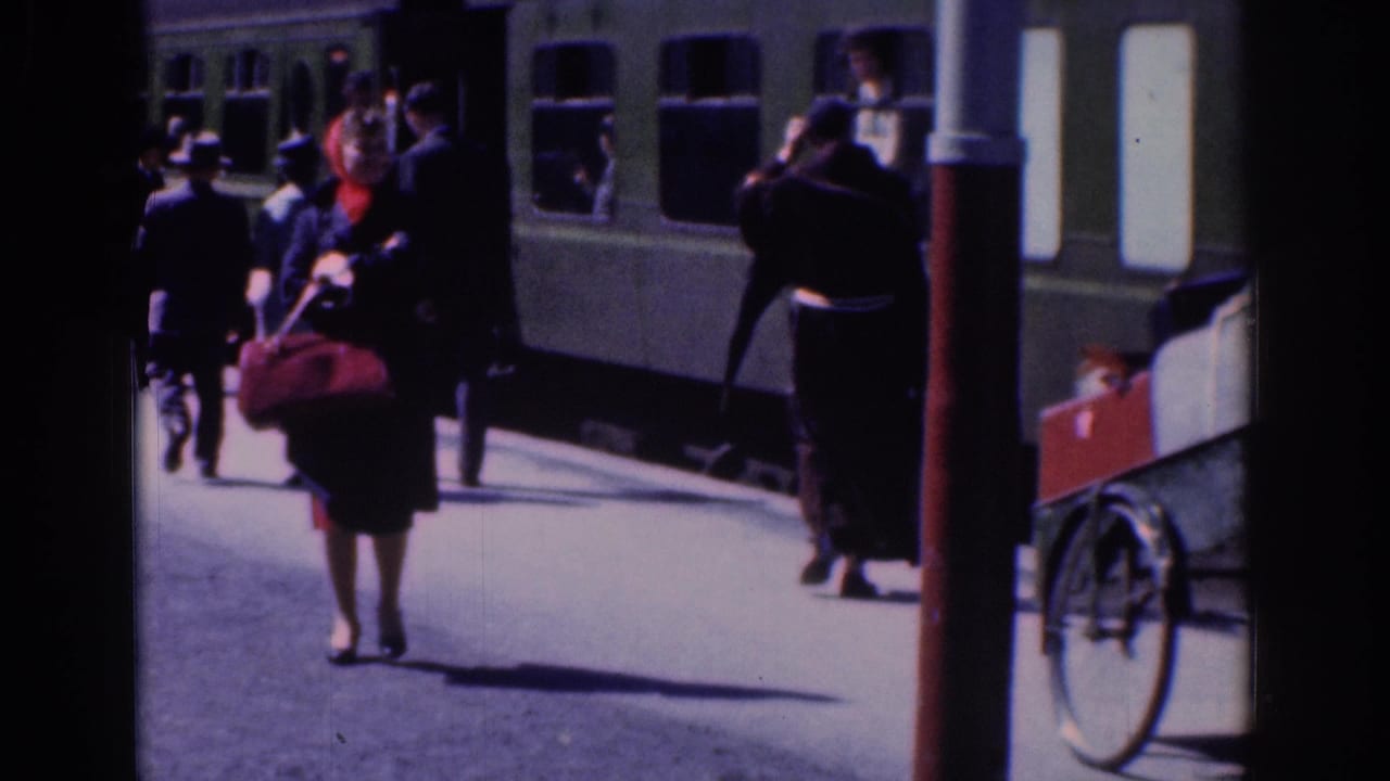 1961 Dublin railway-platform view with passengers near a green train, including a woman carrying a dark bag and wearing a red scarf.