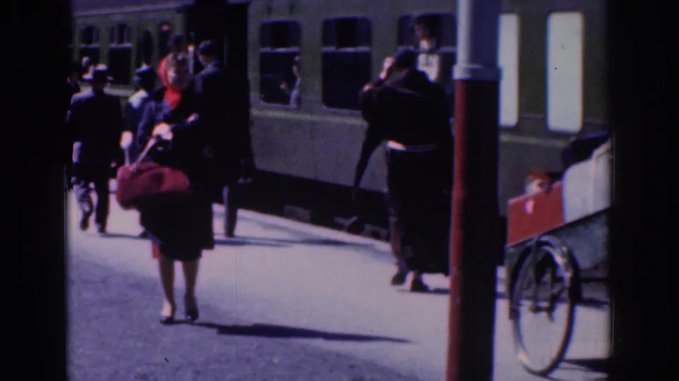 1961 Dublin railway-platform view with passengers near a green train, including a woman carrying a dark bag and wearing a red scarf.