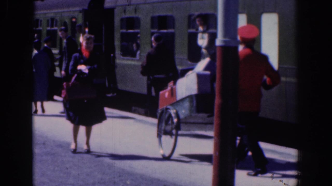 A woman stands on the platform near a luggage cart while a red-uniformed worker walks beside the train.
