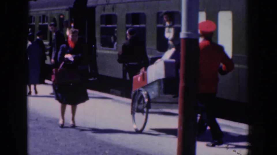 At a Dublin railway platform in 1961, a woman with luggage faces the camera while a red-uniformed rail worker moves near a green carriage.