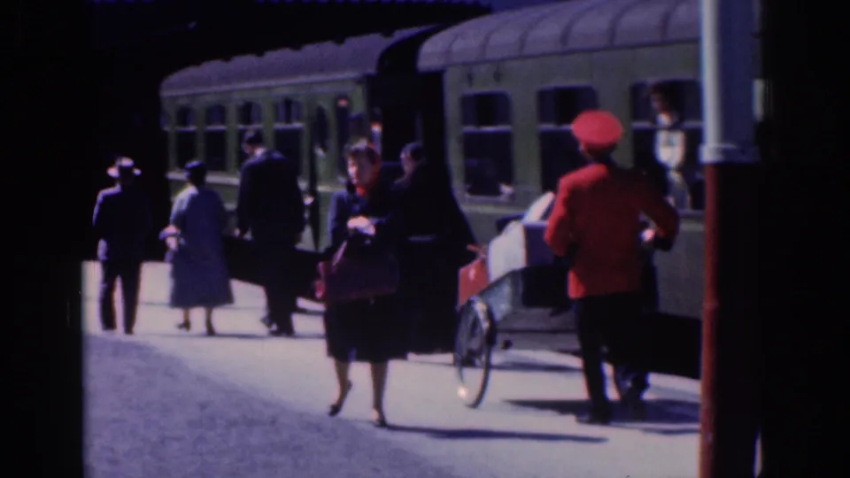 1961 Dublin station-platform frame showing passengers in period clothing, a red-uniformed railway worker, and a luggage cart beside the train.