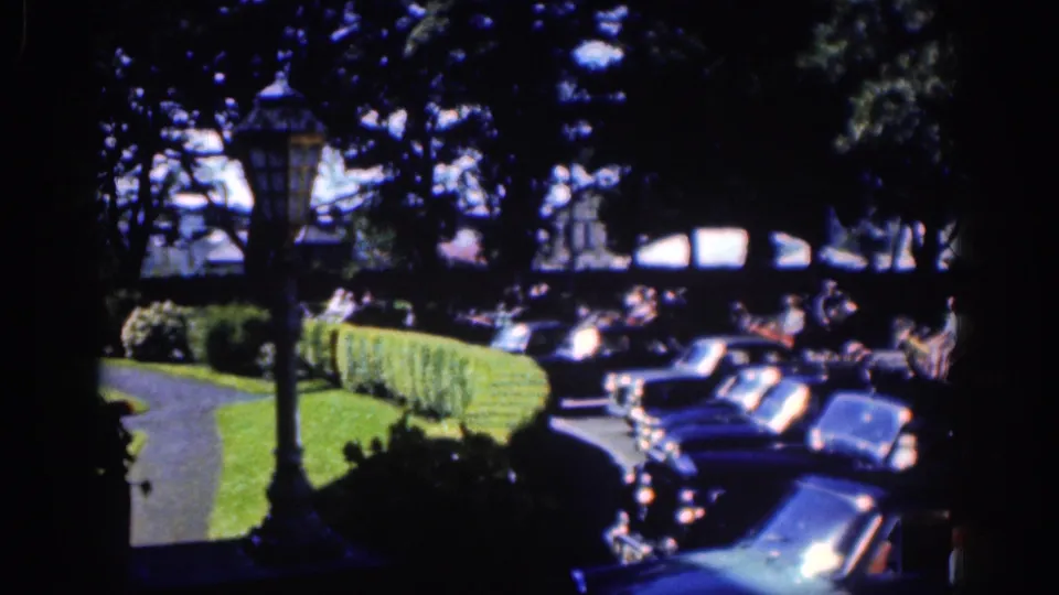1961 Dublin home-movie frame showing a shaded street edge with hedges, lamppost, parked cars, and pedestrians beyond the curb.