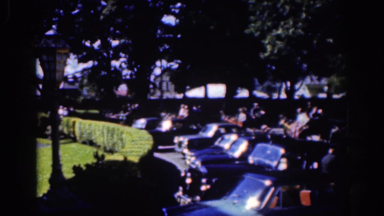 Parked cars line a curb beside hedges while pedestrians gather in the background.
