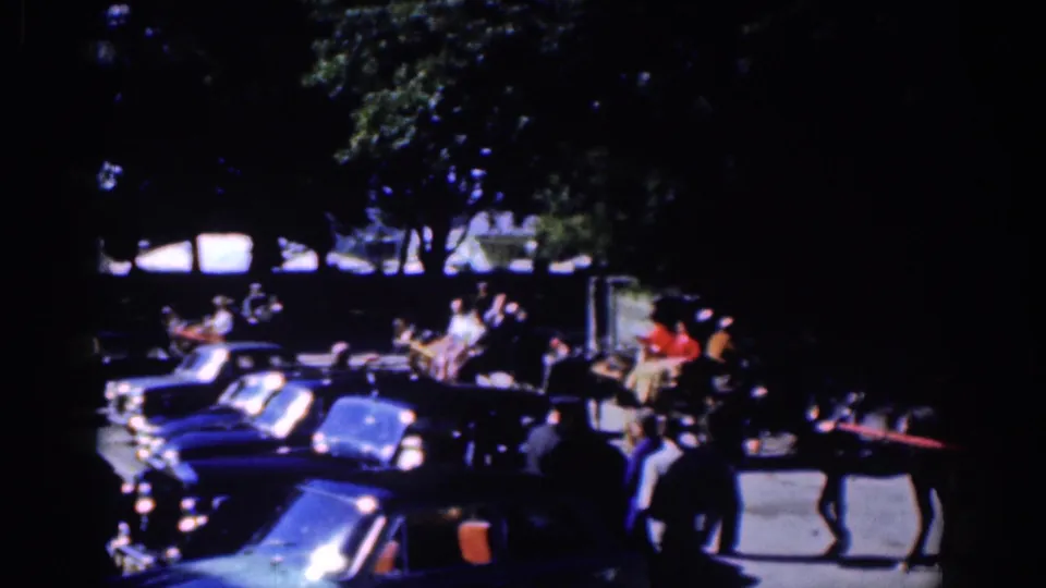 1961 Dublin home-movie frame showing a horse-drawn carriage near parked cars and pedestrians on a tree-shaded street.
