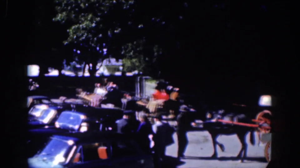 1961 Dublin home-movie frame showing a horse-drawn buggy near parked cars and pedestrians under deep tree shade.