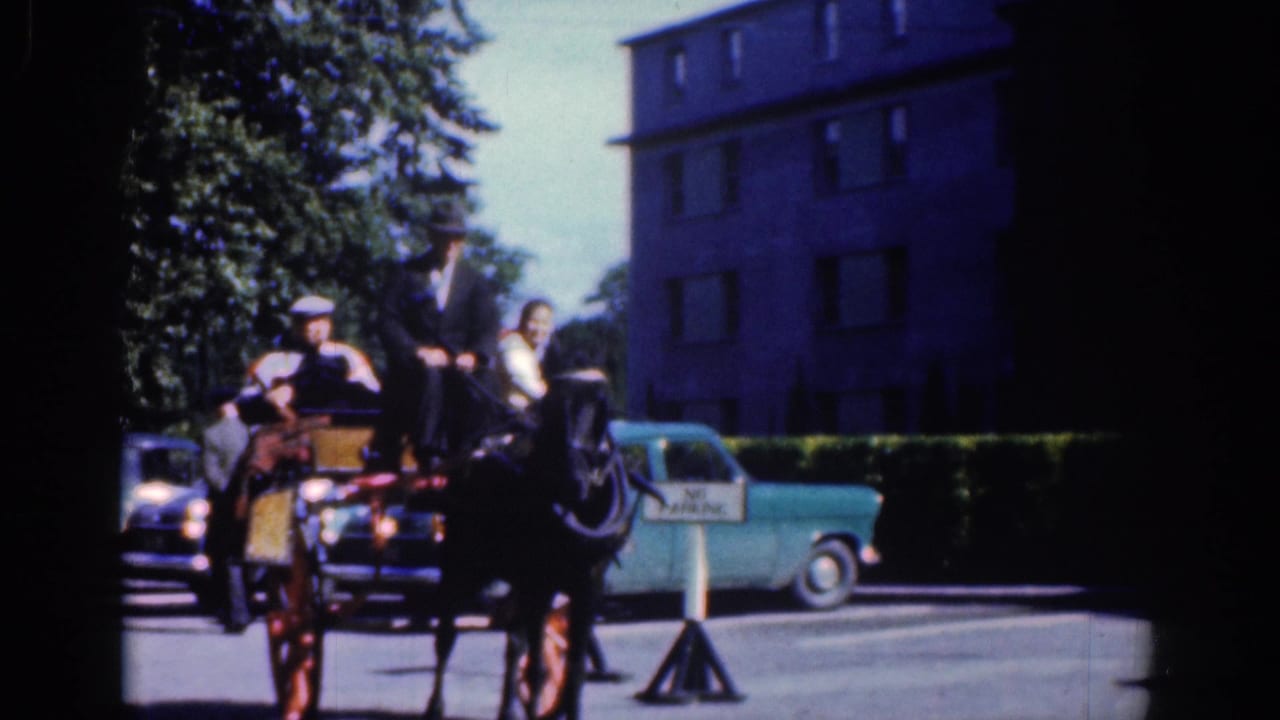 1961 Dublin street view with a horse-drawn carriage, passengers, a suited driver, parked blue cars, trees, and masonry buildings.