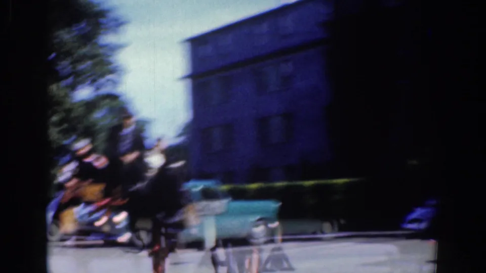 1961 Dublin home-movie frame with motion blur as a horse-drawn carriage passes a pale blue car, hedge, trees, and a large building.