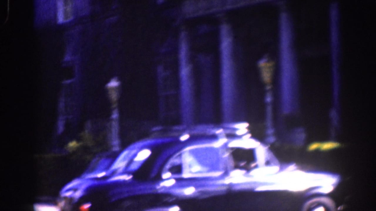 1961 Dublin home-movie frame showing a dark rounded car moving past a shadowed building and street lamps.