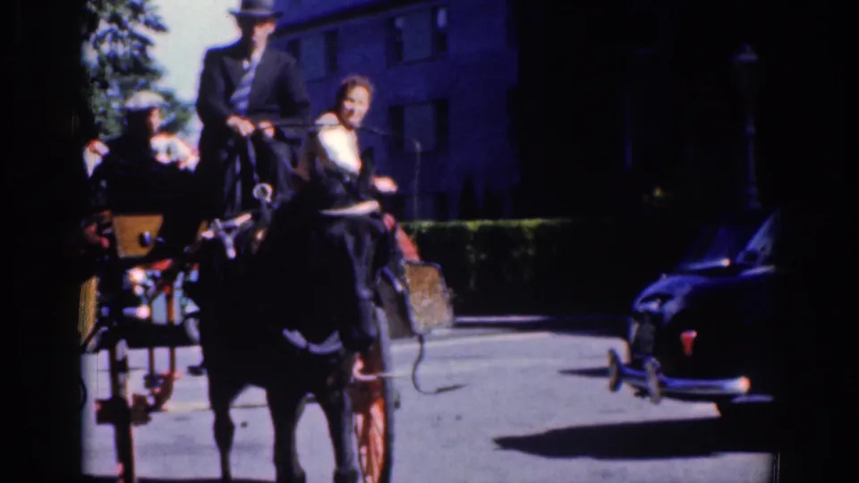 1961 Dublin home-movie frame showing the carriage passing close to camera while a dark car sits or moves at right.