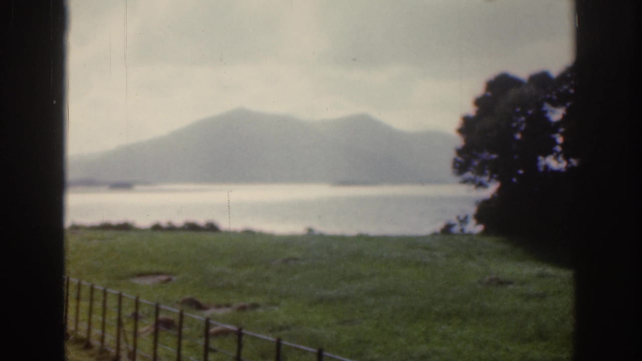 1961 Ireland home-movie frame showing a lake, distant mountains, green field, fence, and dark framing at the edges.