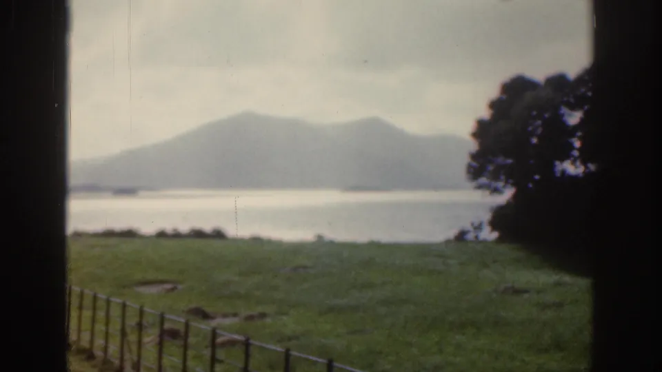 1961 Ireland home-movie frame showing a lake, distant mountains, green field, fence, and dark framing at the edges.