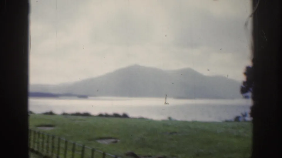 1961 Ireland home-movie frame of a fenced grassy slope leading the eye toward a lake and distant mountains.