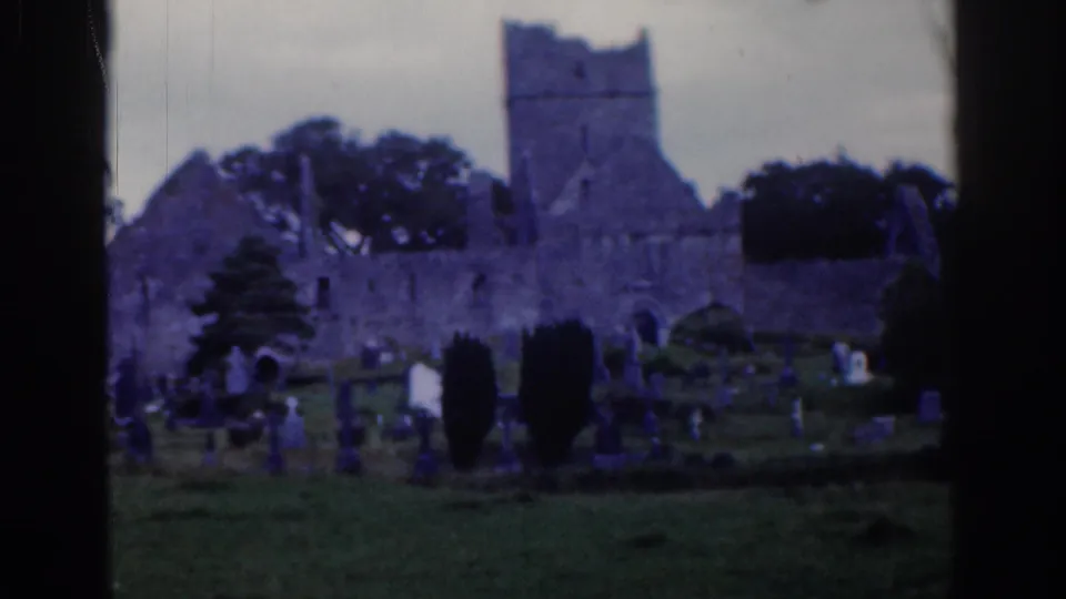 1961 Ireland 8mm frame showing stone castle or abbey ruins beside grave markers, trees, and small visible visitors.