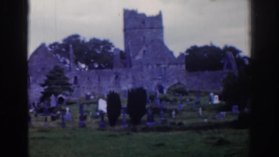 1961 Dublin home-movie frame showing a stone church ruin behind a grassy cemetery with scattered gravestones and trees.