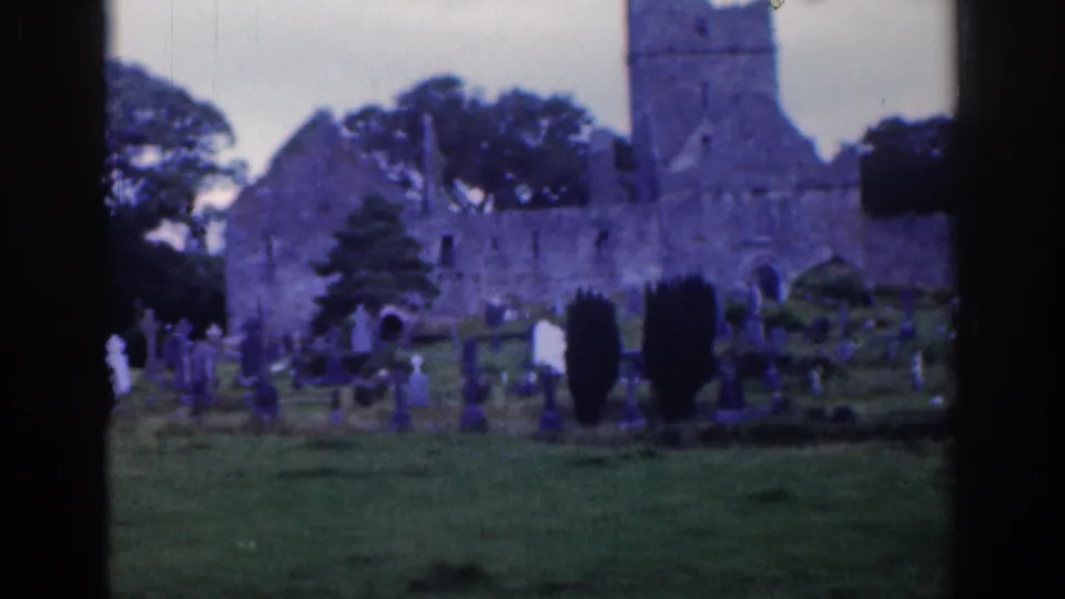 Dublin, 1961, view of a cemetery and old stone church ruin, with trees behind the tower and grave markers across the grass.