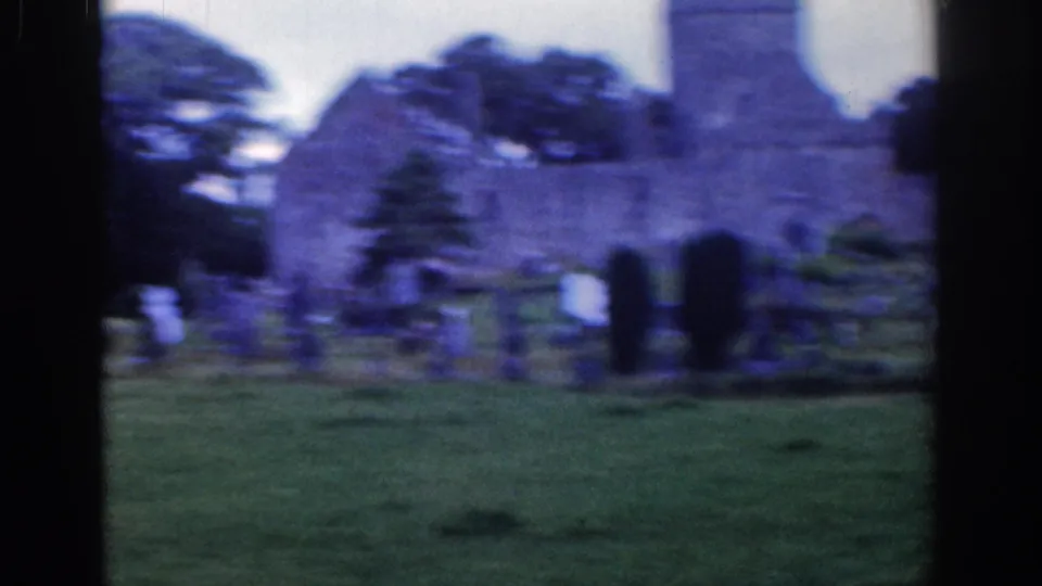 Soft, motion-blurred 1961 Dublin frame of the cemetery lawn, gravestones, trees, and stone church ruin in the background.