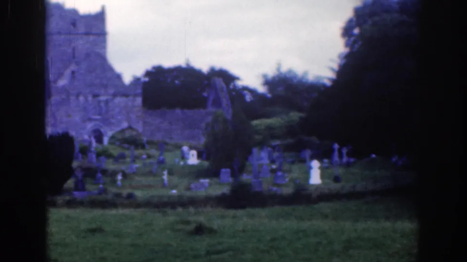 1961 Dublin cemetery view with a ruined stone church, scattered grave markers, dark trees, and cloudy sky on faded color 8mm film.