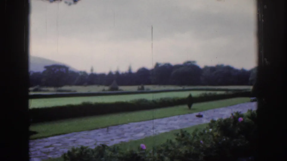 1961 Dublin estate landscape with clipped green lawns, a paved path, hedges, trees, flowers, and distant hills under gray sky.