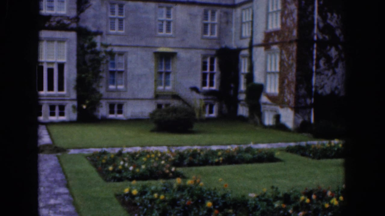 1961 Dublin home-movie frame of a gray stone mansion, clipped lawn, flowerbeds, path, and dark foreground window edge.